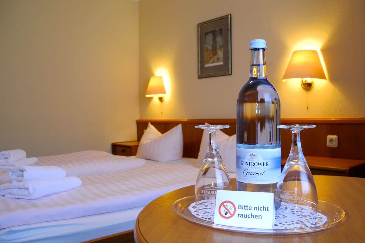Una habitación de hotel con una cama hecha con esmero, una mesita de noche con una botella de agua mineral Güstrower, dos copas de vino y un cartel que indica que no se permite fumar.