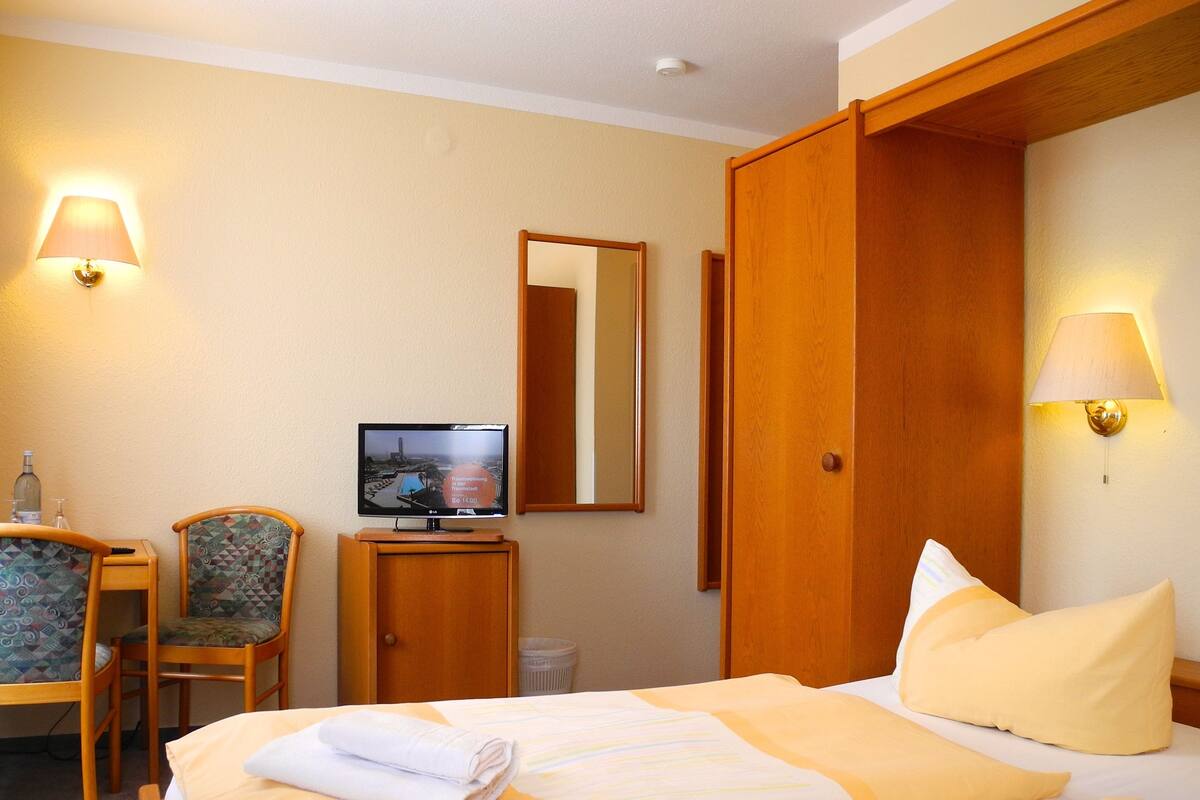 Habitación de hotel con una cama, un televisor sobre un soporte de madera, una silla y un escritorio con una botella y vasos.