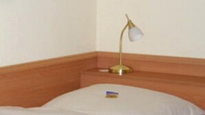 A neatly made bed with white linens, a gold-colored lamp on the nightstand, and a wooden headboard. - Hotel Alfa (Neu-Isenburg)