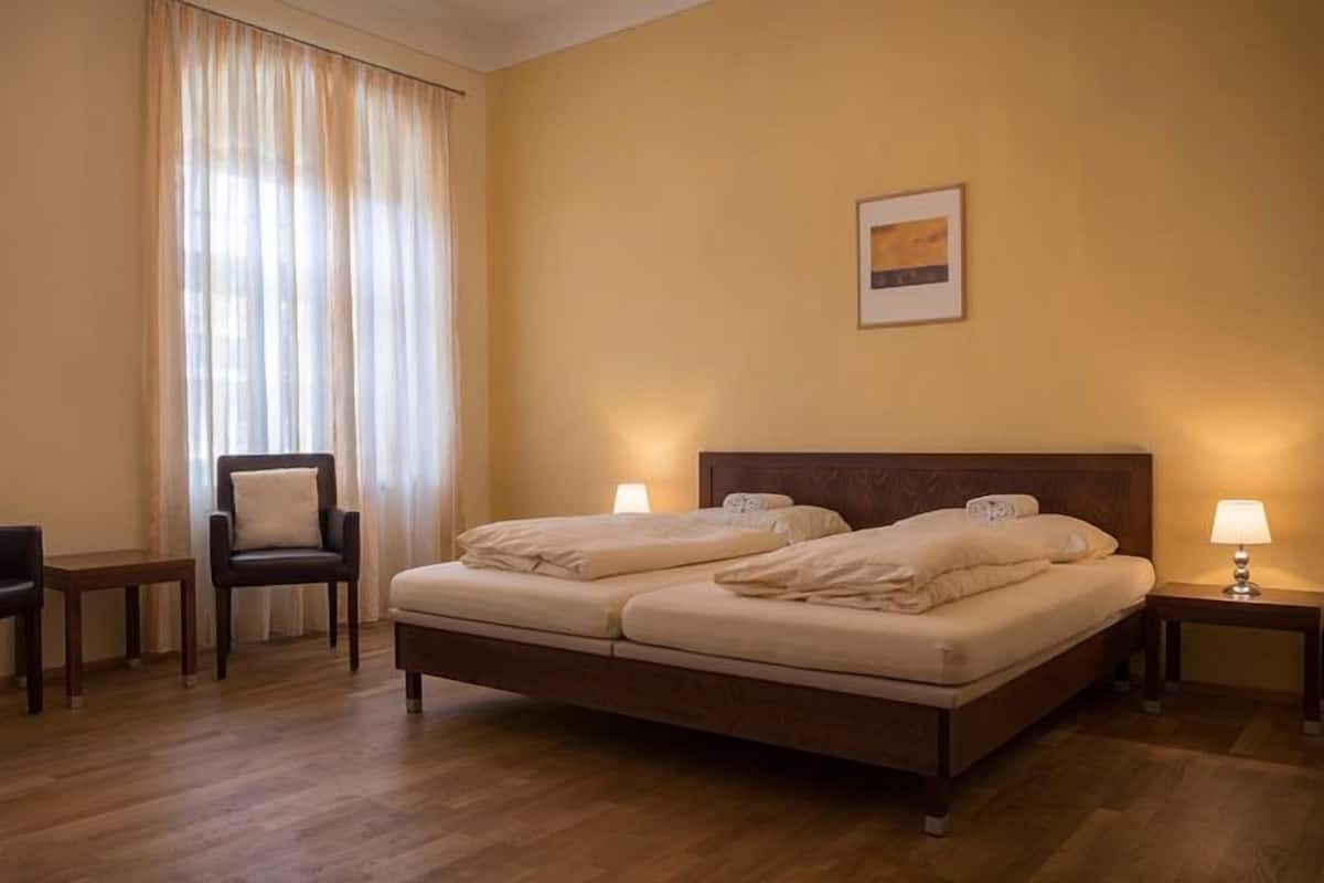 A hotel room with a double bed, two bedside tables, a chair, a small table, and a window with curtains.