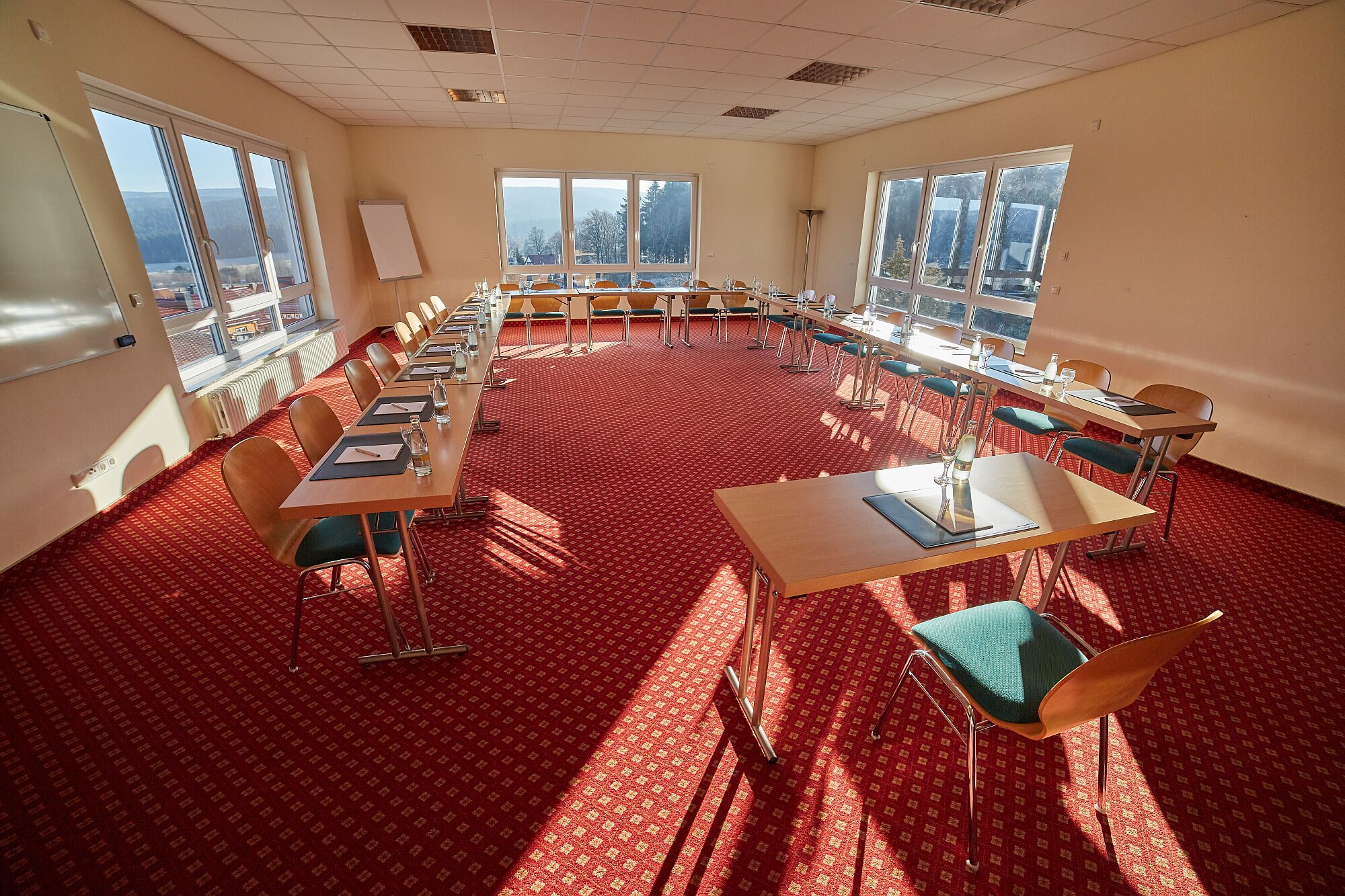A conference room with a red carpet, multiple tables, and chairs, large windows with a view of trees and buildings, and a whiteboard on the wall.