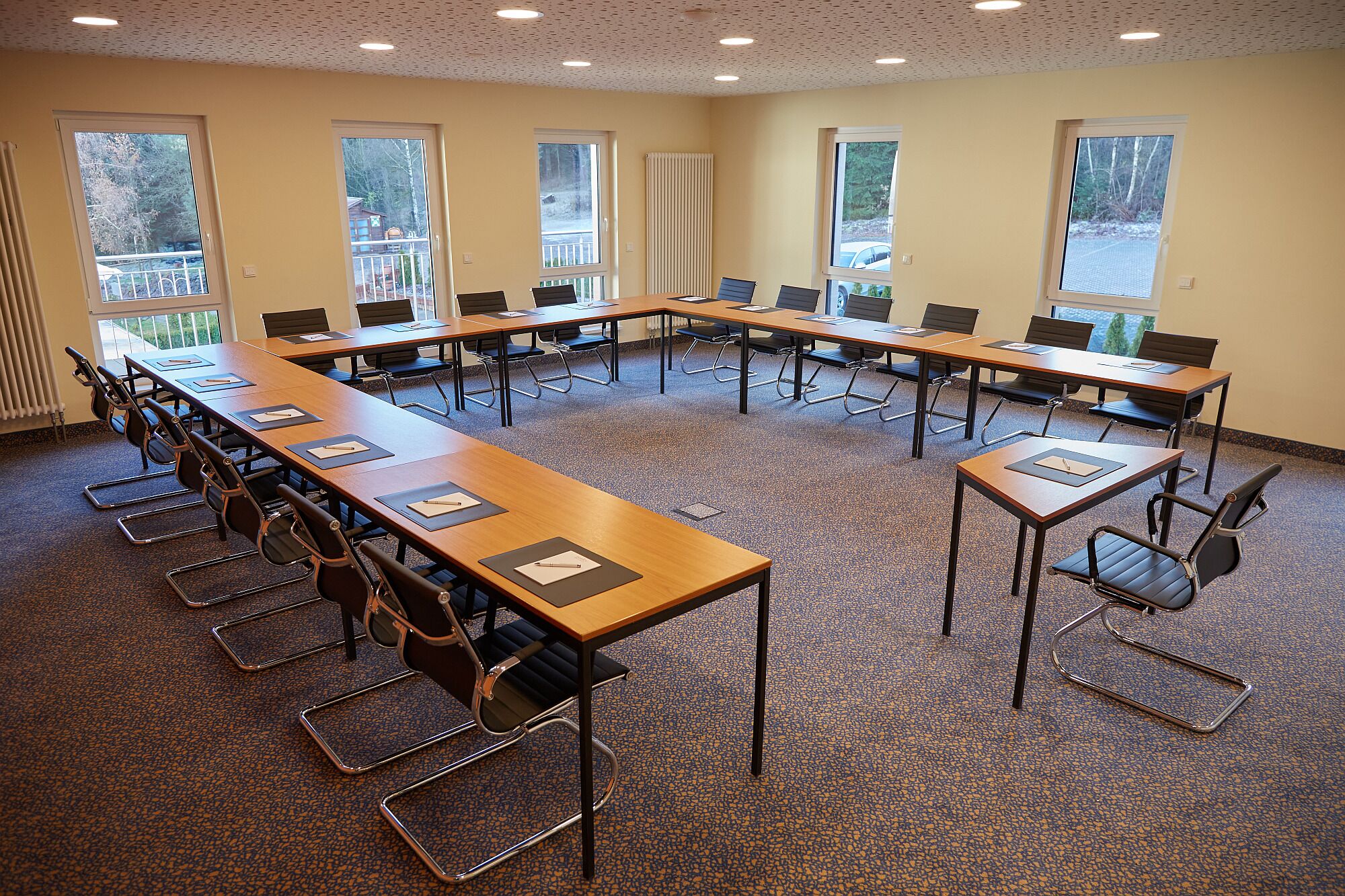 Una sala de conferencias con mesas largas y sillas, ventanas amplias y suelo alfombrado.