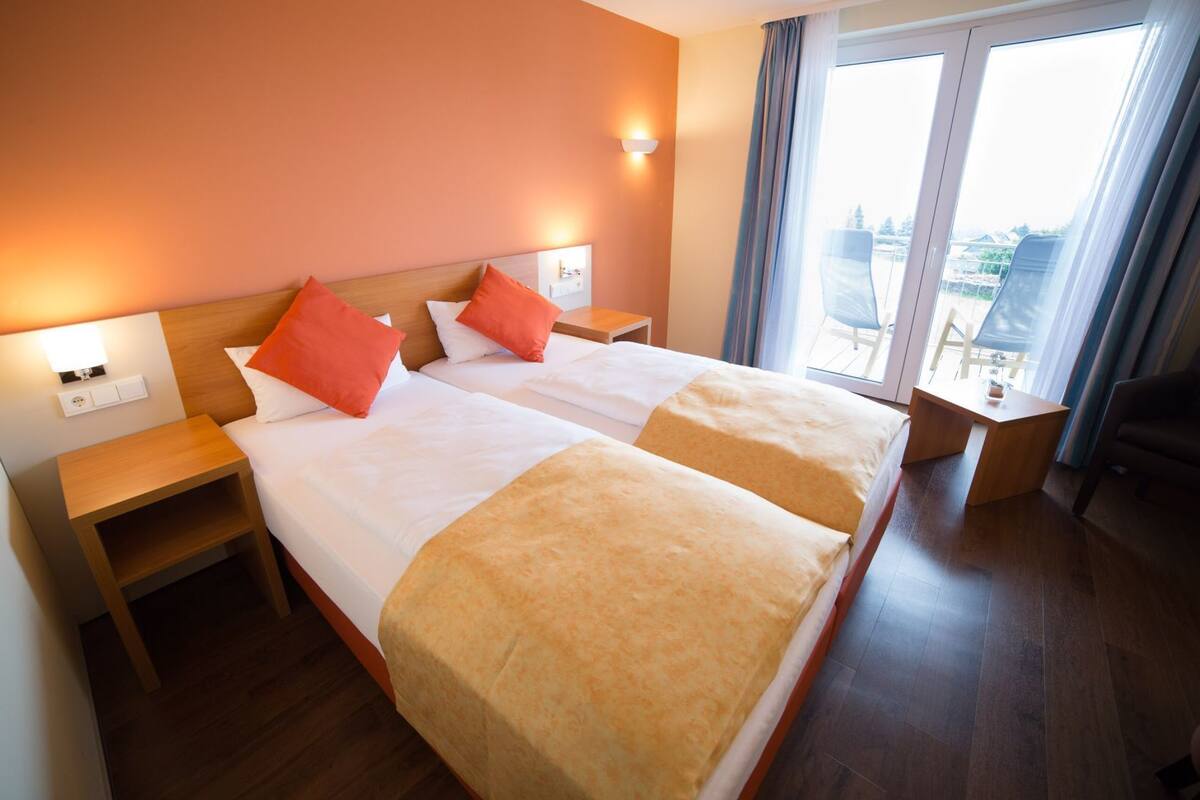 Habitación de hotel con dos camas, ropa de cama anaranjada y blanca, cabecero de madera, mesitas de noche y vista de la ciudad a través de ventanas amplias.