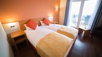 Habitación de hotel con dos camas, ropa de cama anaranjada y blanca, cabecero de madera, mesitas de noche y vista de la ciudad a través de ventanas amplias.