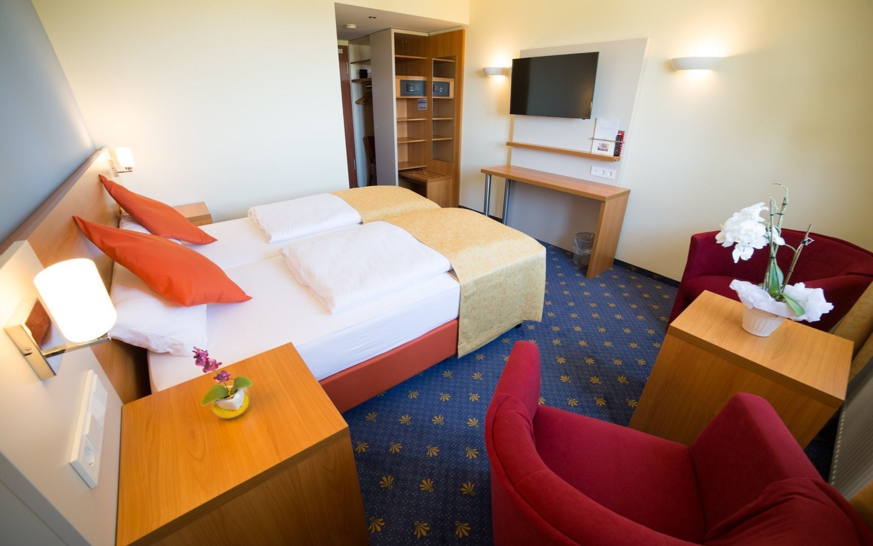 A hotel room with a bed, a desk, a TV, a chair, and a small table with a flower vase.