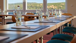 Un salón de conferencias con mesas dispuestas para reuniones, equipado con botellas de agua y vasos, y amplias ventanas que ofrecen vistas a un paisaje boscoso.