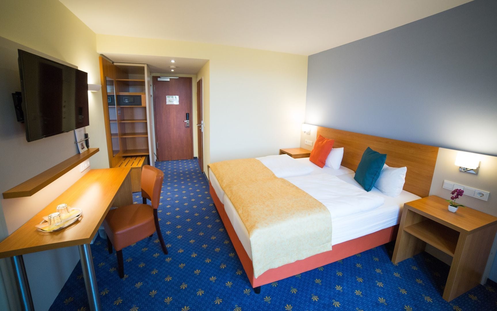 A hotel room with a bed, desk, chair, and a TV on the wall.