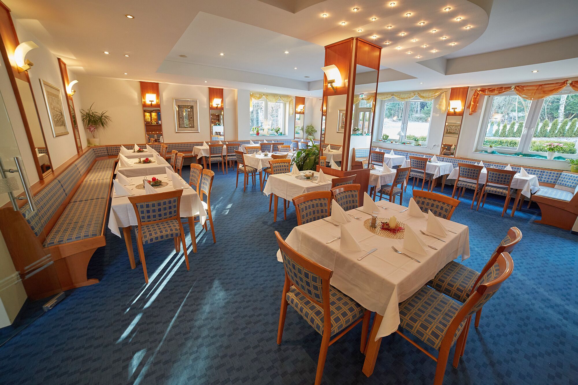 A restaurant with multiple tables and chairs, a blue carpeted floor, and large windows with views of greenery.