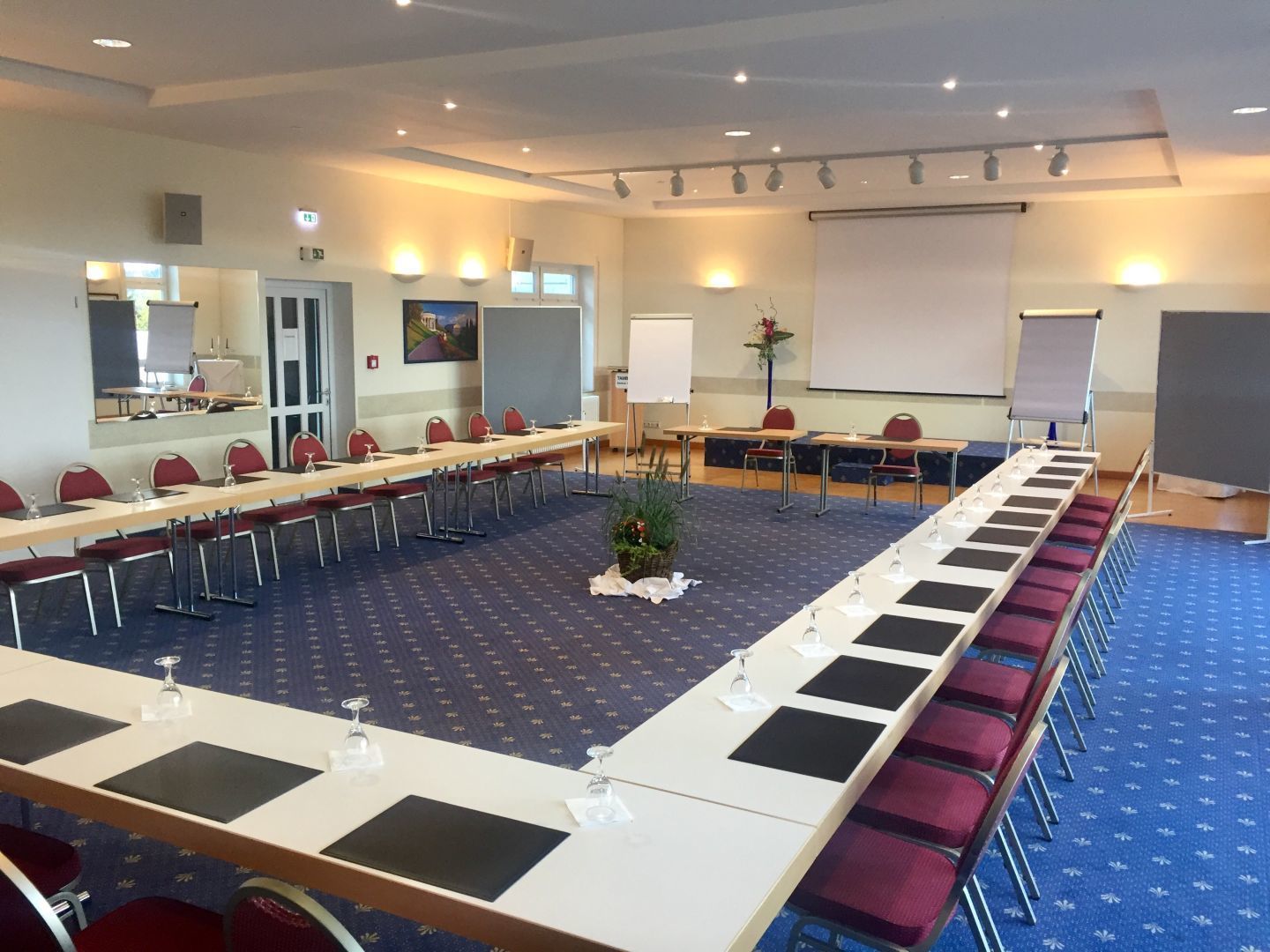 Un salón de conferencias con largas mesas, sillas, una pantalla de proyector y una pizarra.