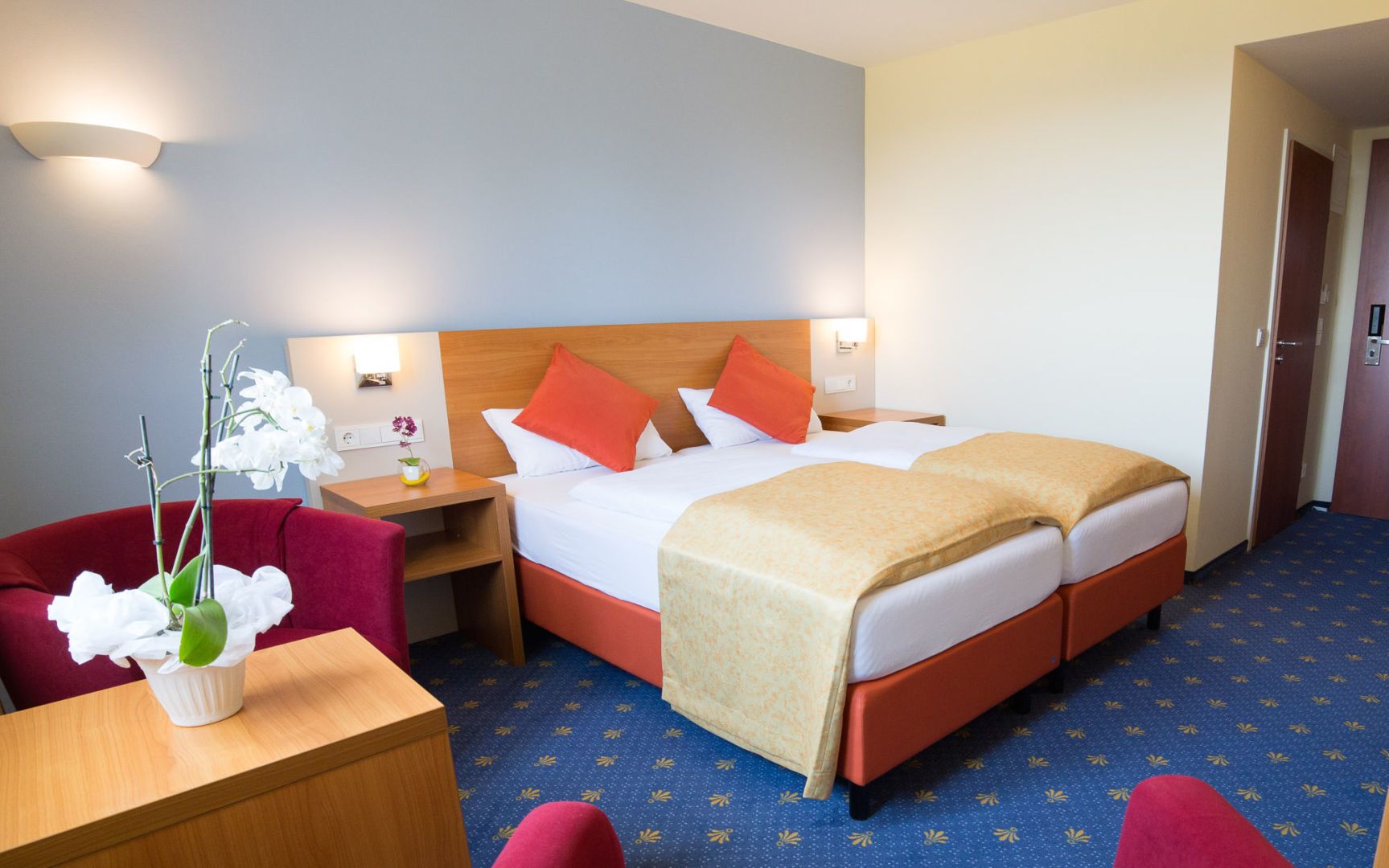 Hotelkamer met een tweepersoonsbed, twee nachtkastjes, een bureau met stoel, een rode fauteuil en een kleine potplant.