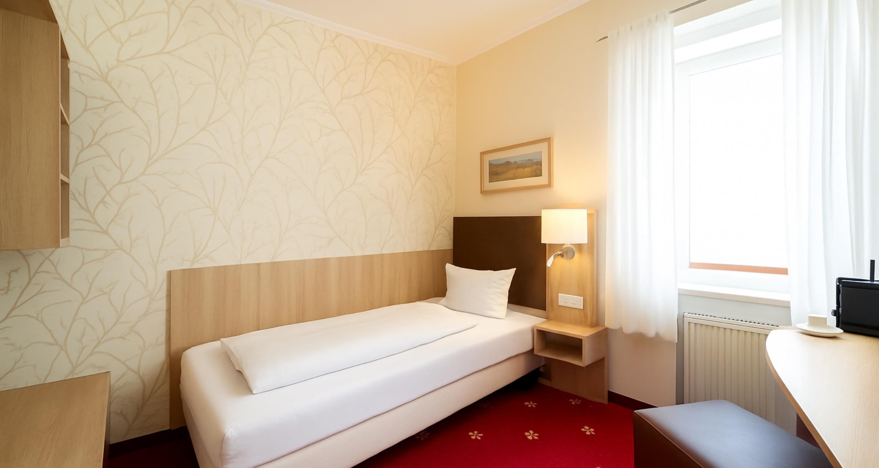 A hotel room with a single bed, a bedside table with a lamp, a small bench, a window with curtains, and a painting on the wall.