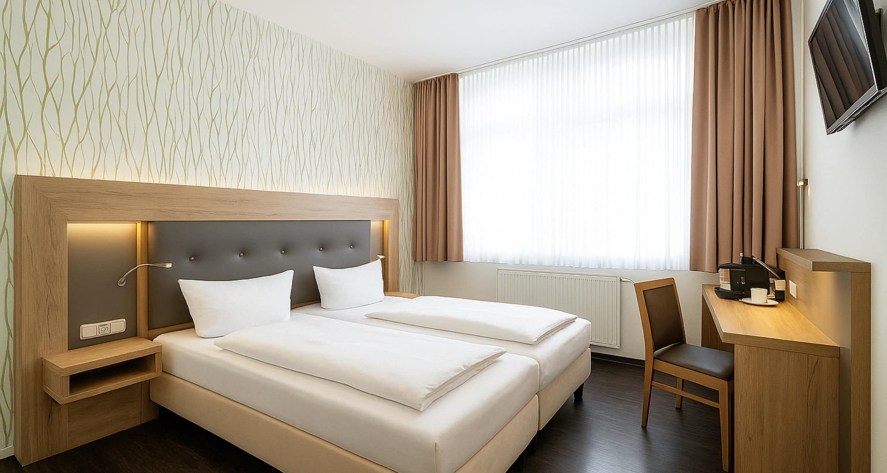 Habitació d'hotel amb dos llits, una taula, una cadira, un televisor i una finestra amb cortines.