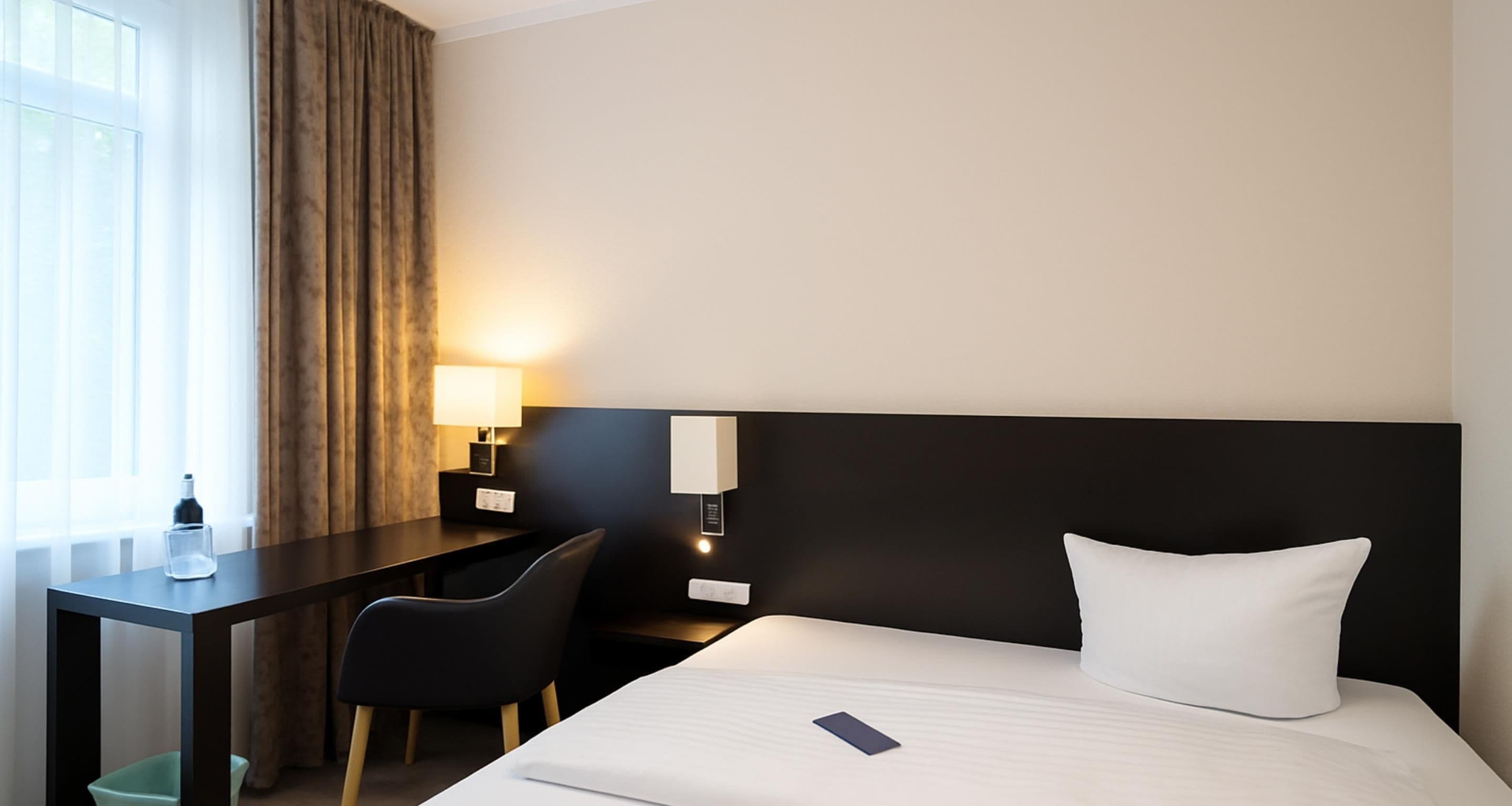 Une chambre d’hôtel avec un lit, un bureau, une chaise, une lampe et une fenêtre avec des rideaux.