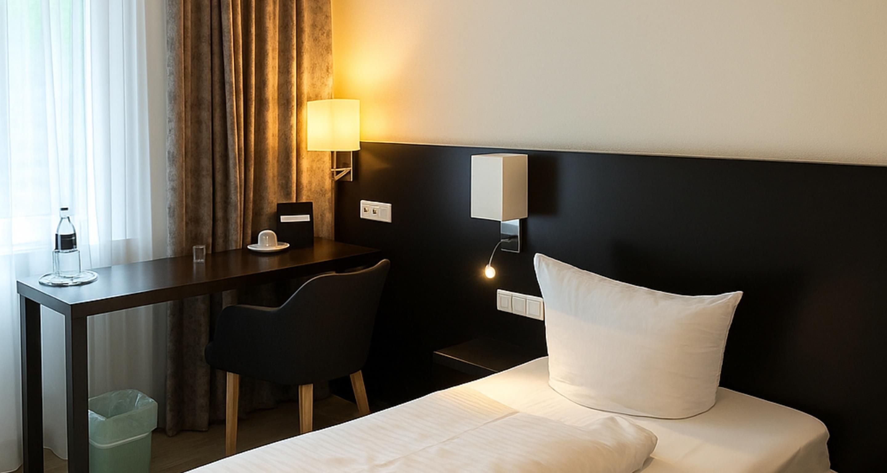 A hotel room with a bed, a desk, a chair, a lamp, and a trash bin.