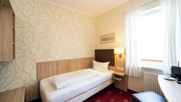 A hotel room with a single bed, a nightstand with a lamp, a window with curtains, and a radiator.