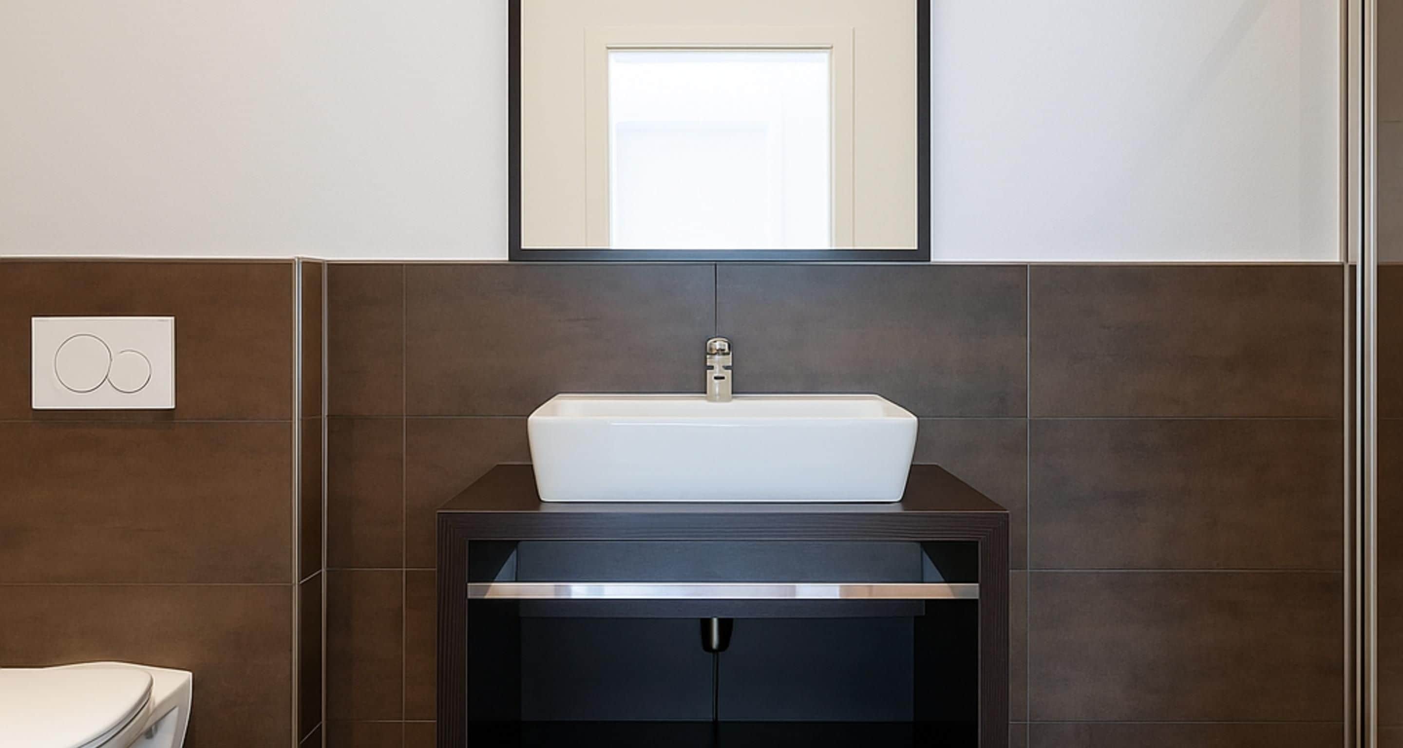 Un baño con un lavabo blanco, baldosas oscuras, un espejo sobre el lavabo y un inodoro a la izquierda.
