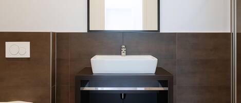 Un baño con un lavabo blanco, baldosas oscuras, un espejo sobre el lavabo y un inodoro a la izquierda.