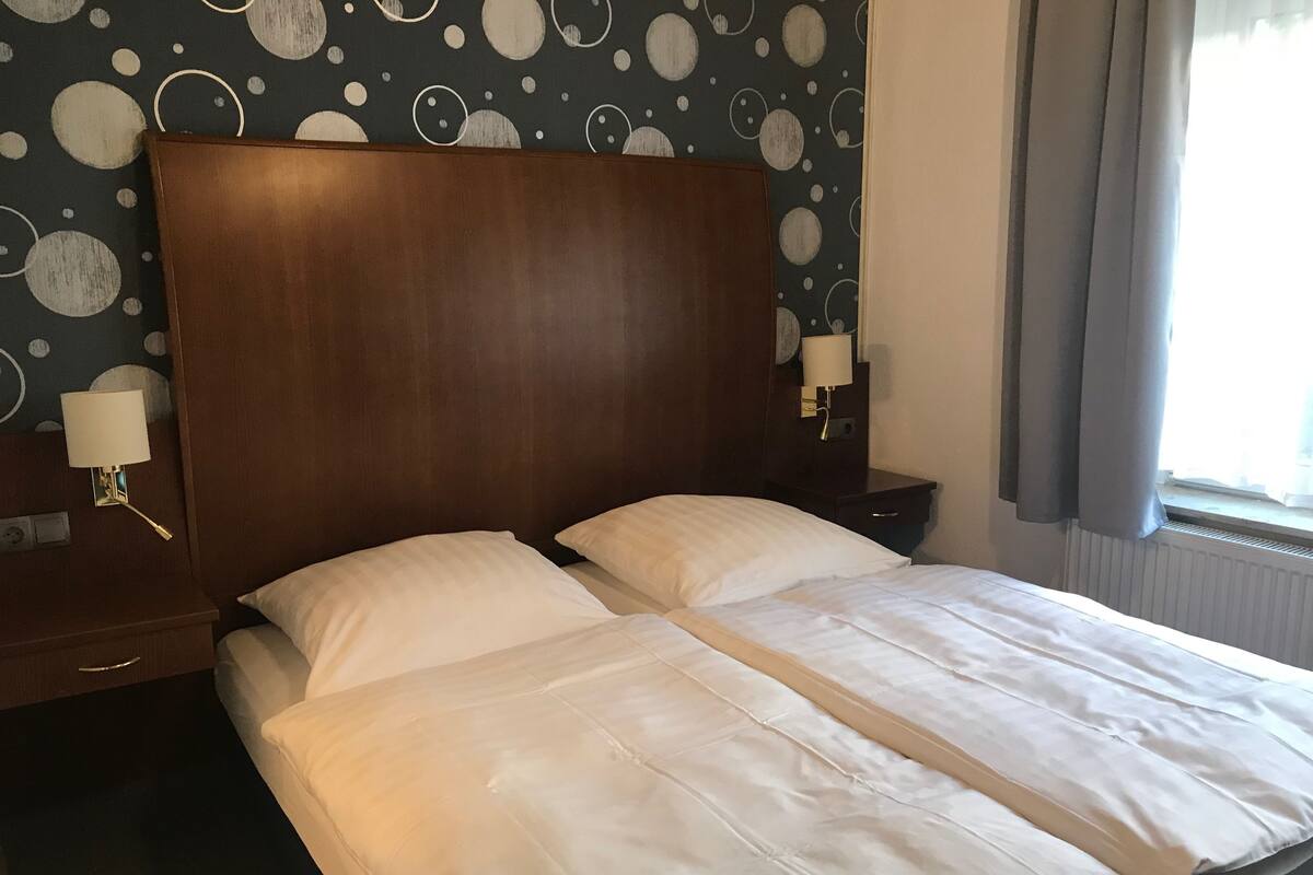 A double bed with white linens, a wooden headboard, and a wall with a circular pattern. There are two bedside tables with lamps.