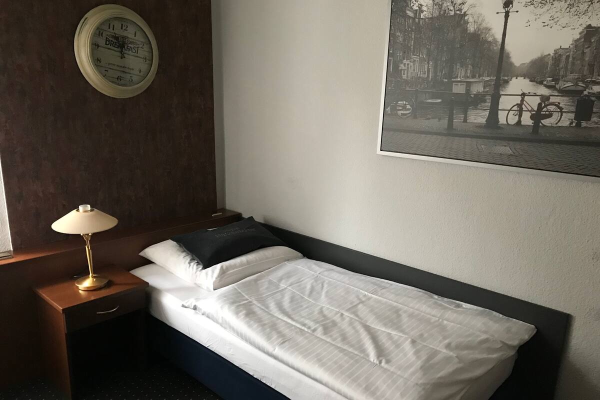 A hotel room with a single bed, a bedside table with a lamp, a wall clock, and a framed picture of a canal scene.