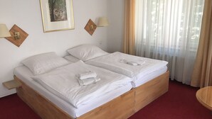 Una cama doble con sábanas y toallas blancas, cabecera de madera, mesita de noche pequeña y una lámpara de pared.