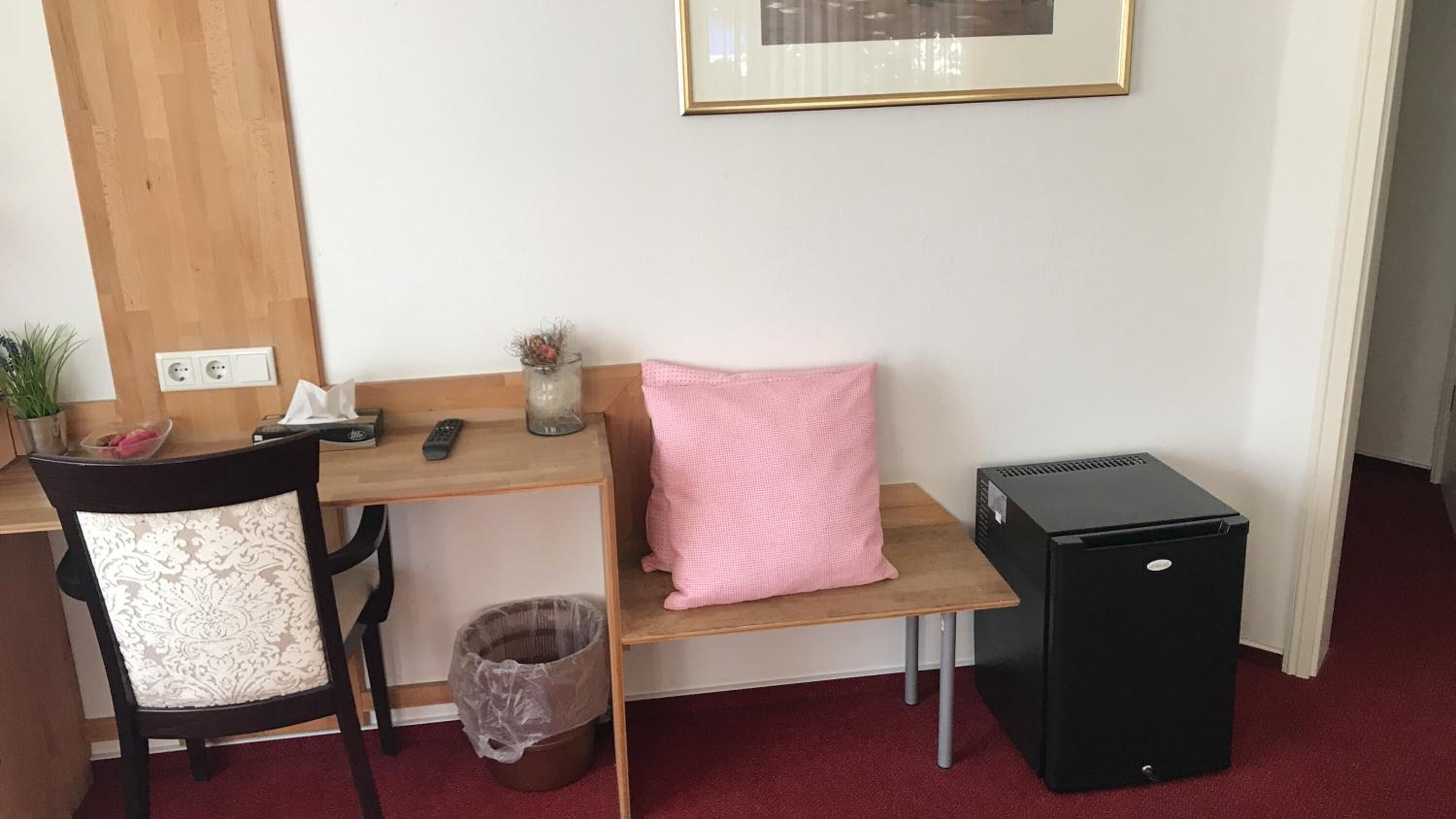 A small, tidy room with a wooden desk, a black computer, a chair with a white cushion, a pink pillow, a small trash bin, and a framed picture on the wall.