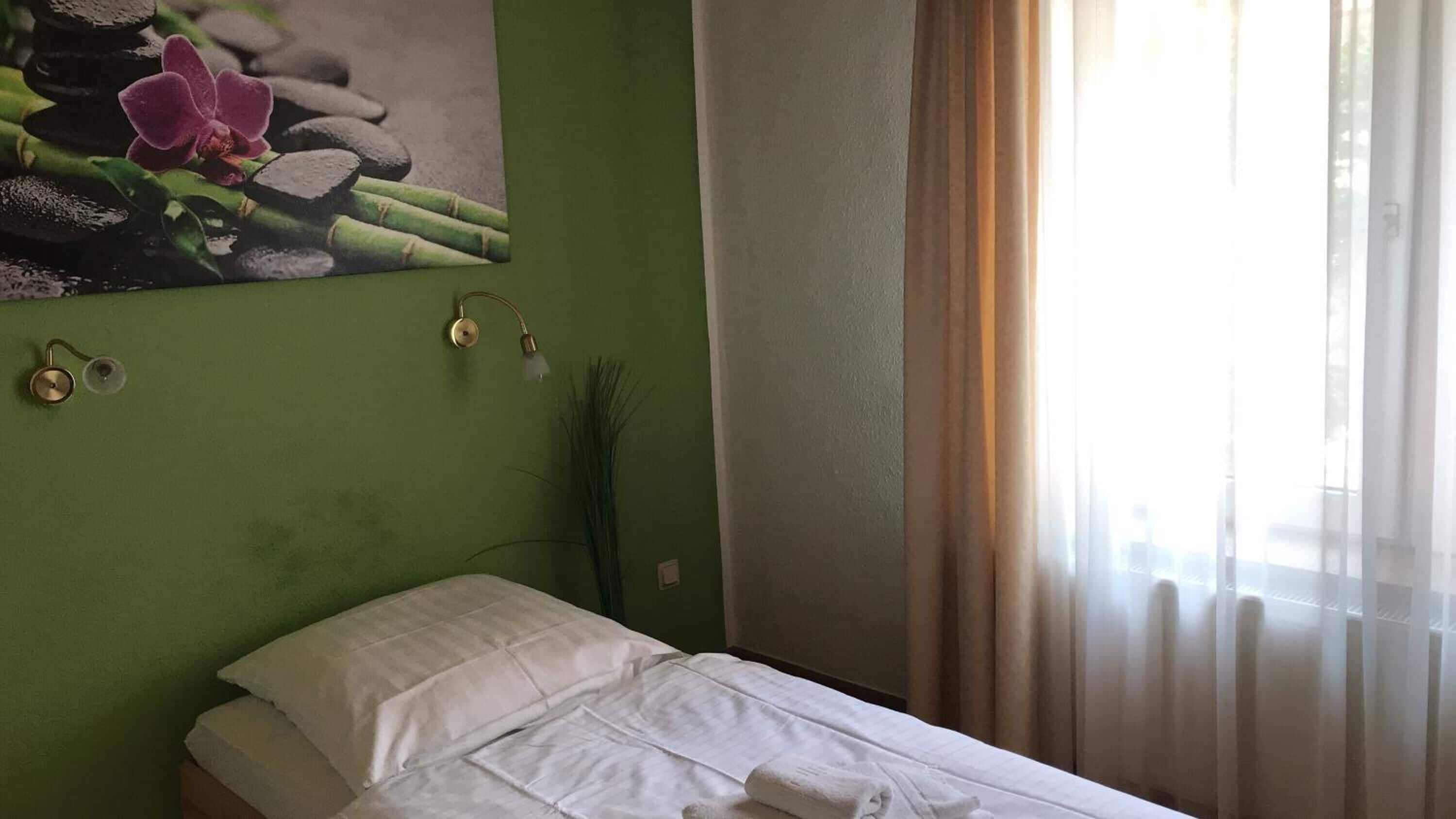 Quarto de hotel com uma cama, uma parede verde, um quadro com pedras e flores, e uma janela com cortinas.