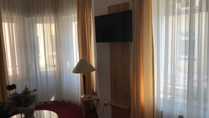Una habitación con cortinas translúcidas, un televisor de pantalla plana montado, una mesa con lámpara y un jarrón con flores.