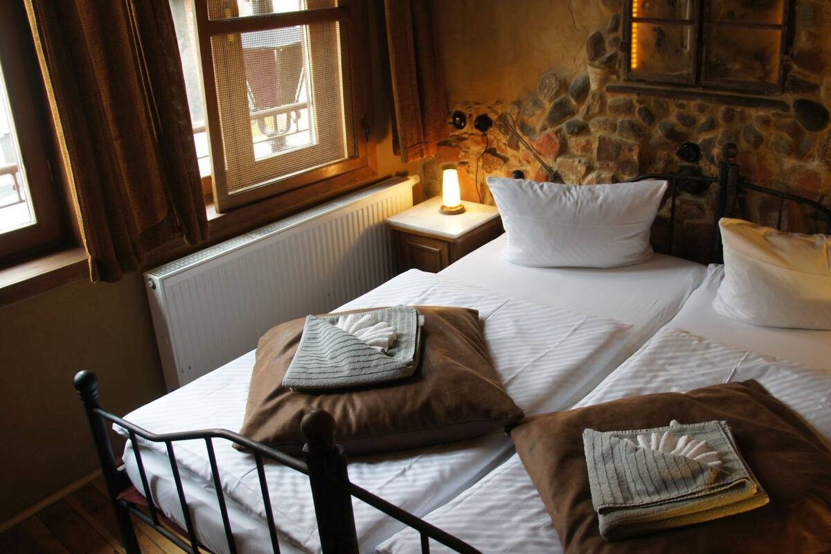 Una cama con sábanas blancas, dos almohadas marrones y una mesita de noche con una lámpara.