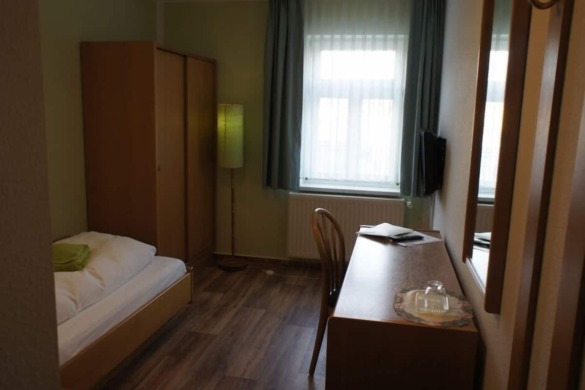Una habitación pequeña y ordenada, con cama, escritorio, silla, lámpara y ventana con cortinas.