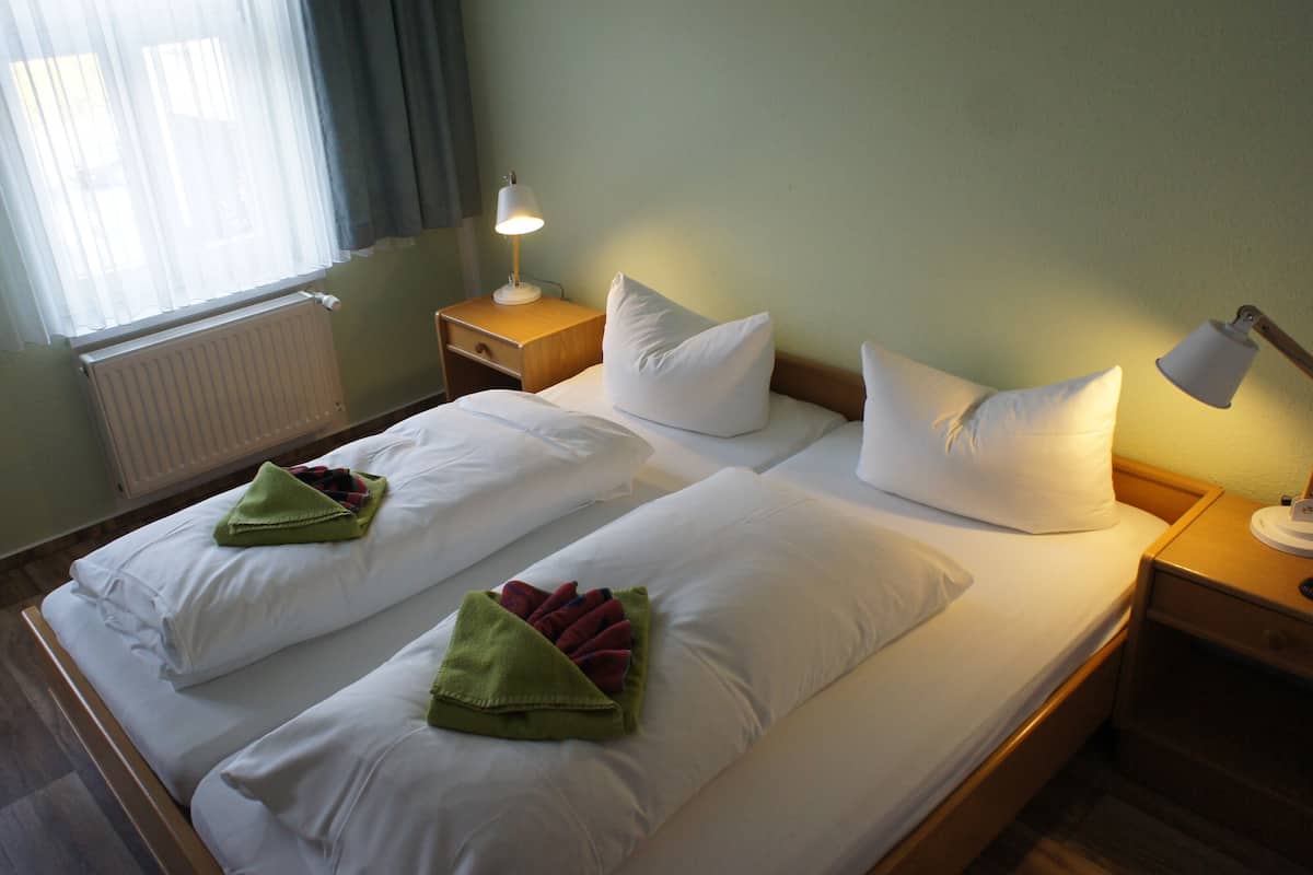 Habitación de hotel con dos camas, ropa de cama blanca, una mesita de noche con lámpara, una ventana con cortinas y un radiador.