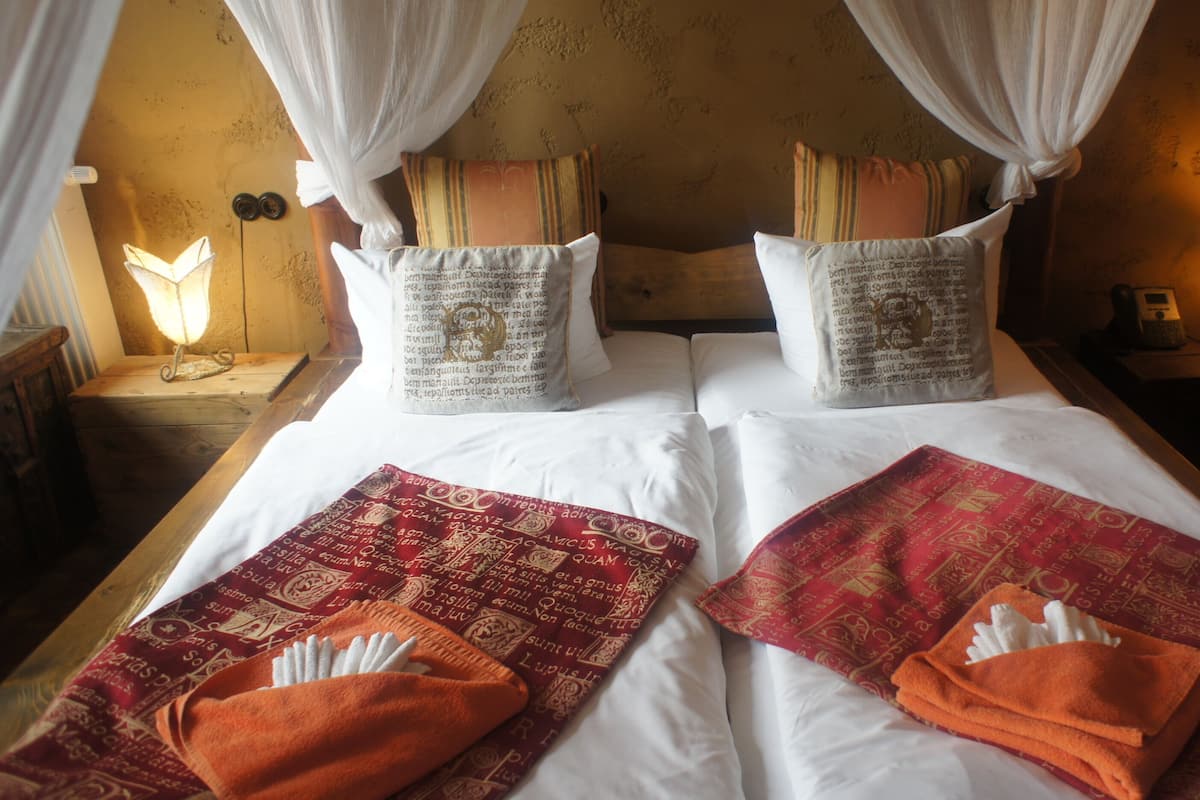 Una cama doble con ropa blanca, dos almohadas con texto y una colcha roja y naranja.