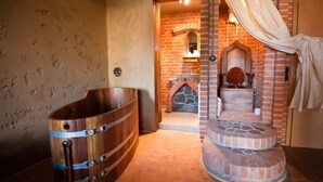 A rustic bathroom with a wooden bathtub, a stone fireplace, and a brick wall.