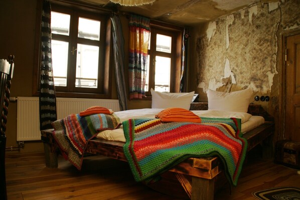 A rustic room with a wooden bed, colorful blankets, a window with curtains, and a stone wall.