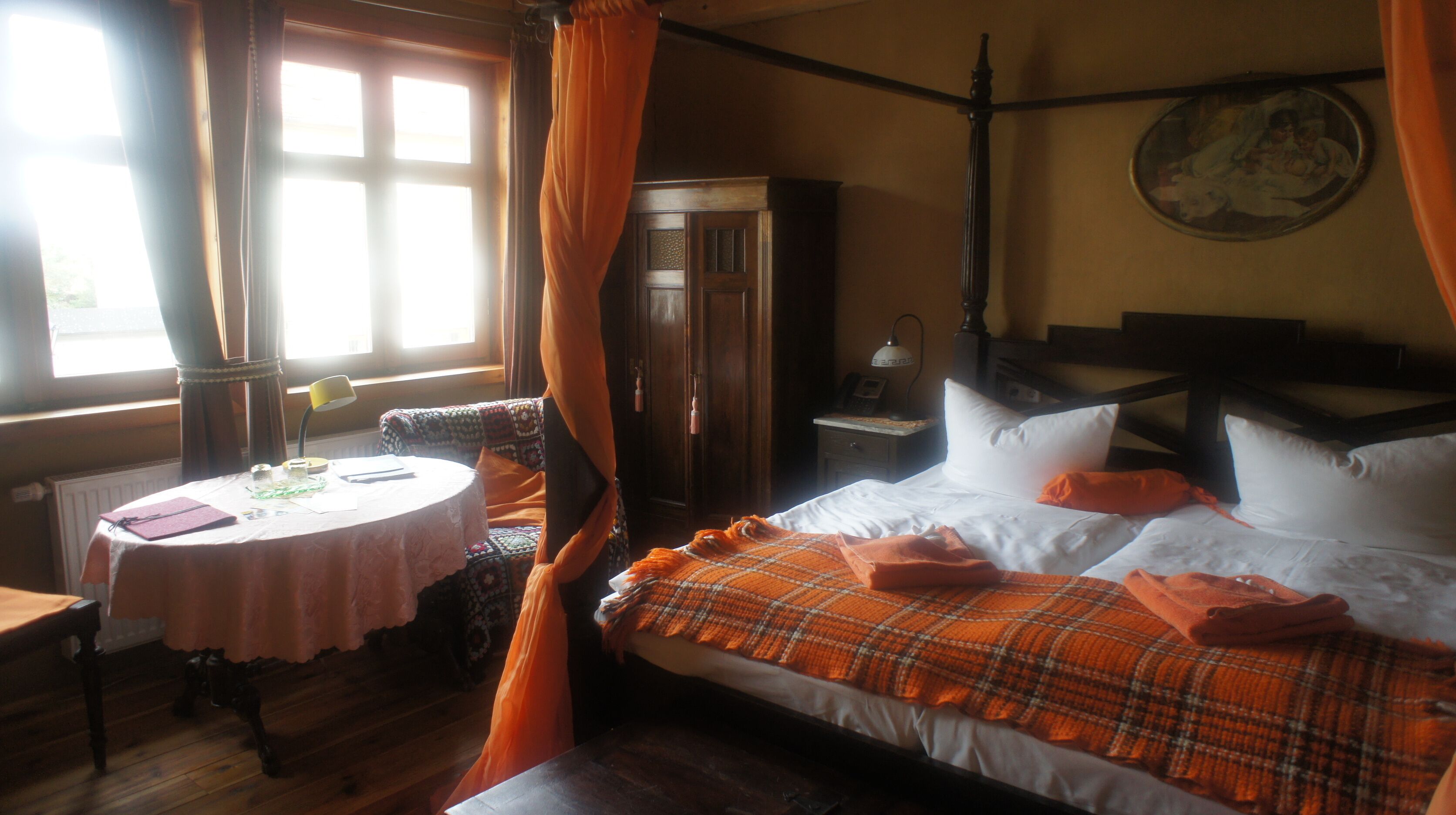 Een kamer met een bed, een tafel en een stoel. Er is een raam met gordijnen, een lamp en een schilderij aan de muur.