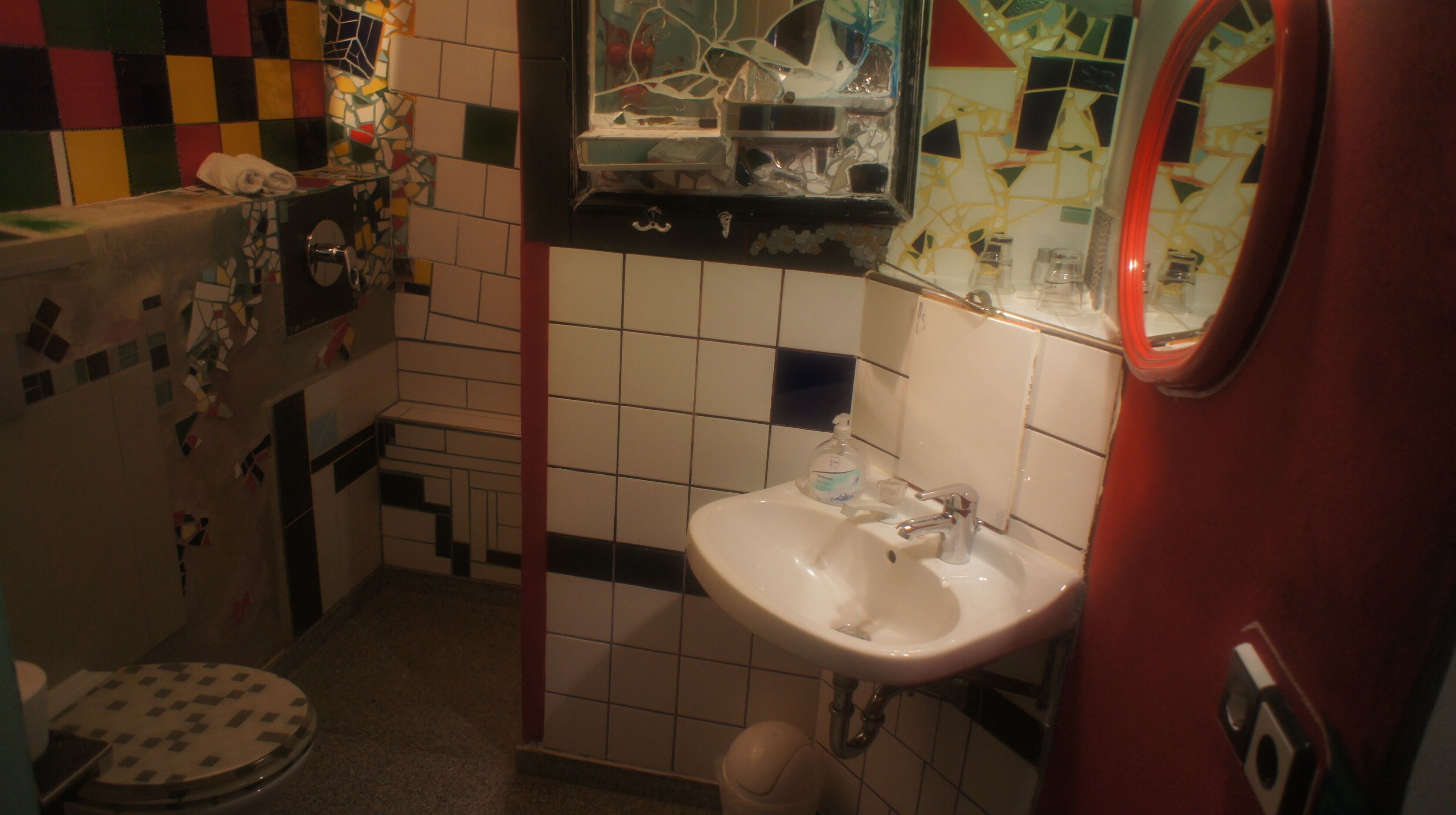 A bathroom with a white sink, a mirror, and a toilet. The walls are tiled with a mix of white and colored tiles.