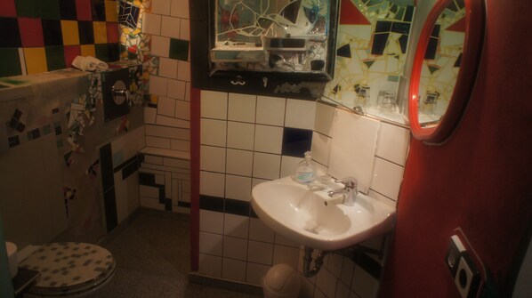A bathroom with a white sink, a mirror, and a toilet. The walls are tiled with a mix of white and colored tiles. - Exempel Schlafstuben Und Quartier Langer Hals (Tangermünde)