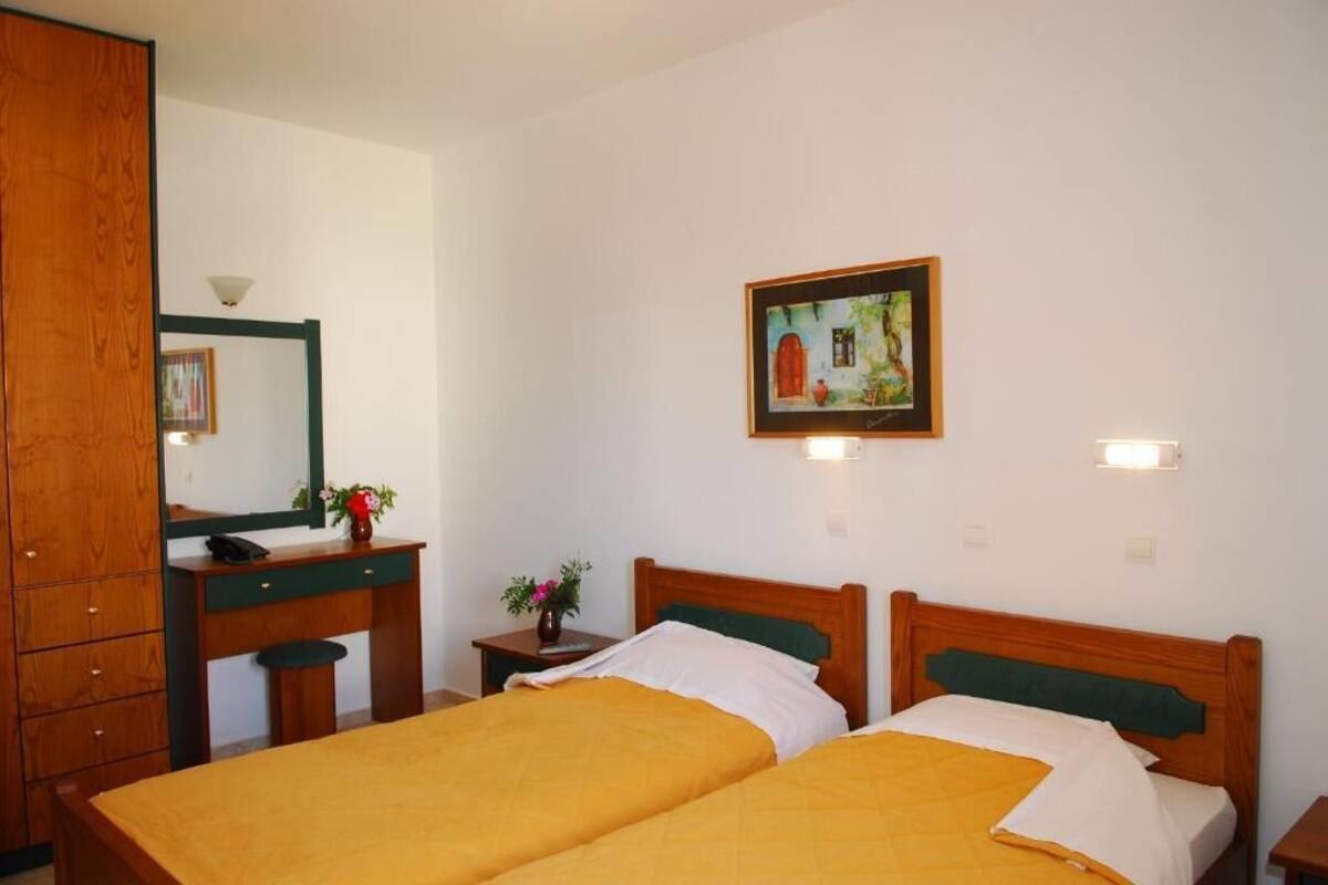 A hotel room with two beds, a wooden wardrobe, a desk with a mirror, a small table with a vase, and a painting on the wall.
