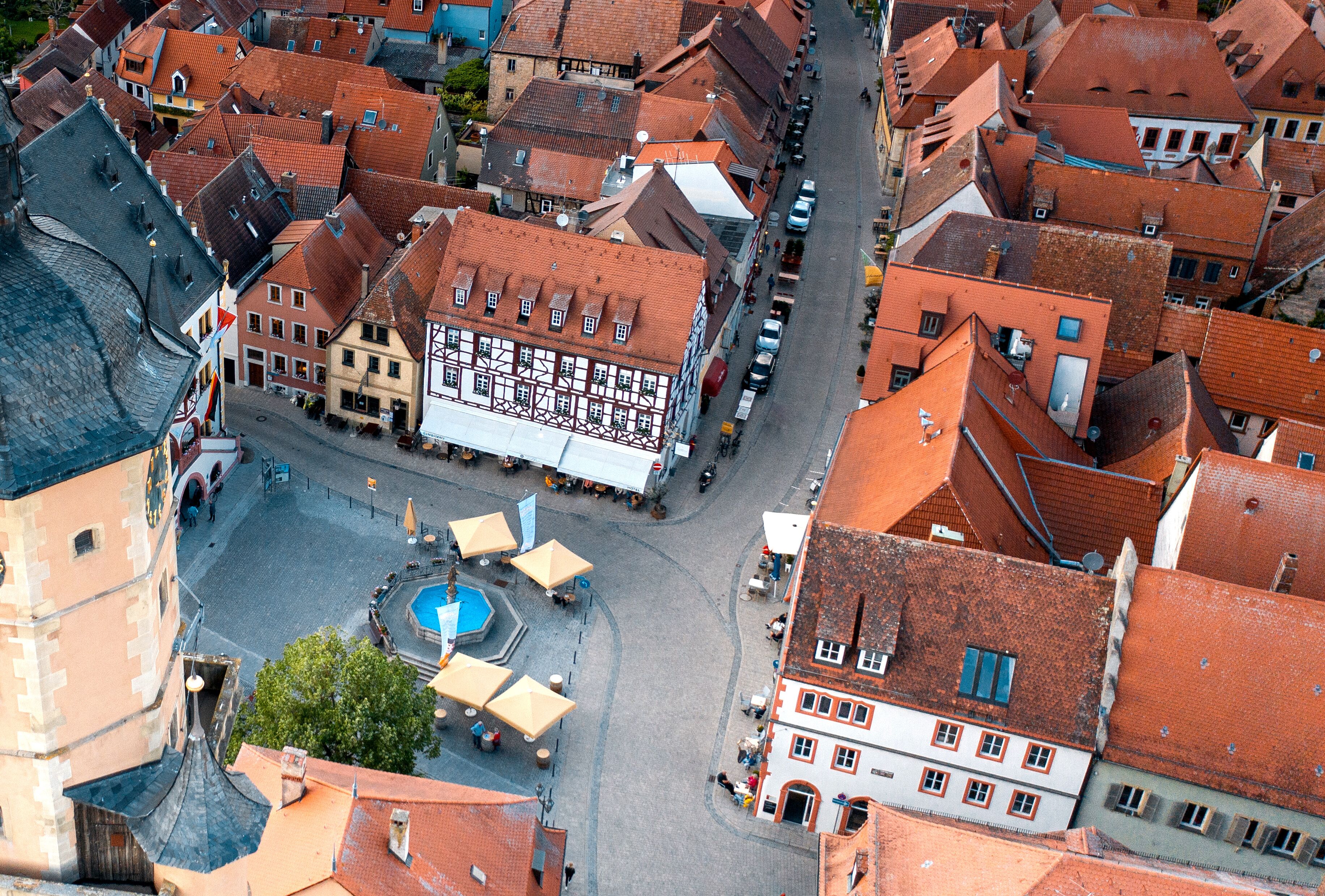 An aerial view of a historic town square with traditional buildings, a fountain, and outdoor seating areas.
