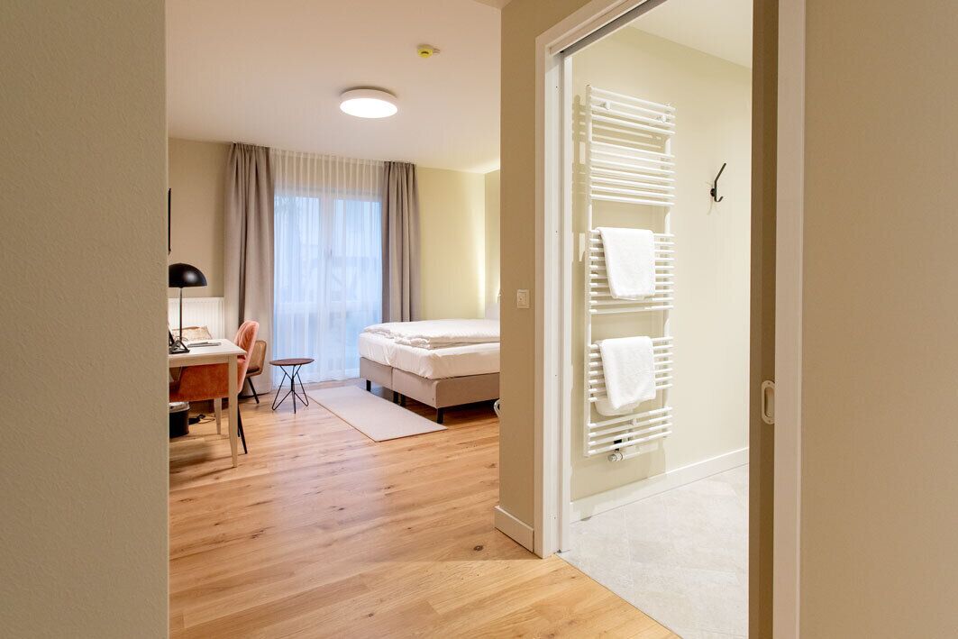 A hotel room with a wooden floor, a bed, a desk with a chair, a lamp, and a window with curtains.