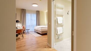 A hotel room with a wooden floor, a bed, a desk with a chair, a lamp, and a window with curtains.