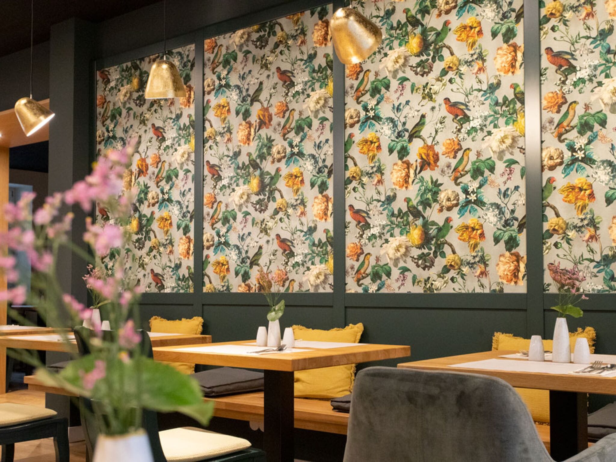 A restaurant interior with wooden tables, yellow cushions, and a floral wallpaper featuring birds and flowers.