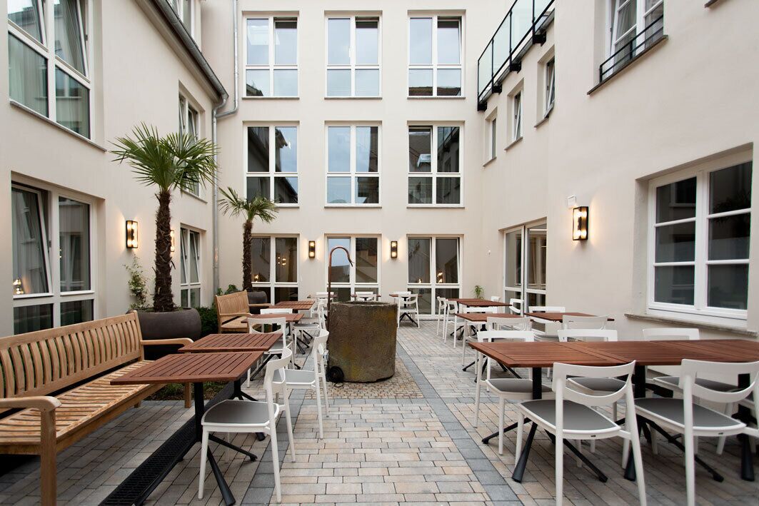 An outdoor patio with wooden tables and chairs, a central stone feature, and a building with multiple windows in the background.