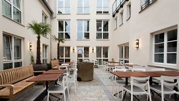 An outdoor patio with wooden tables and chairs, a central stone feature, and a building with multiple windows in the background.