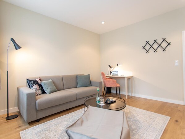 Una sala de estar moderna con un sofá gris, una mesa redonda para café, un escritorio con una silla y una lámpara de pie.