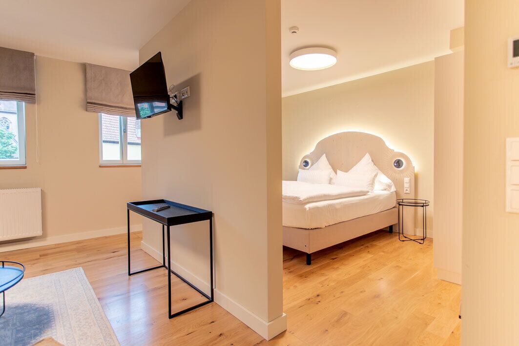 A modern bedroom with a bed, a flat-screen TV mounted on the wall, a small table, and a window with blinds.