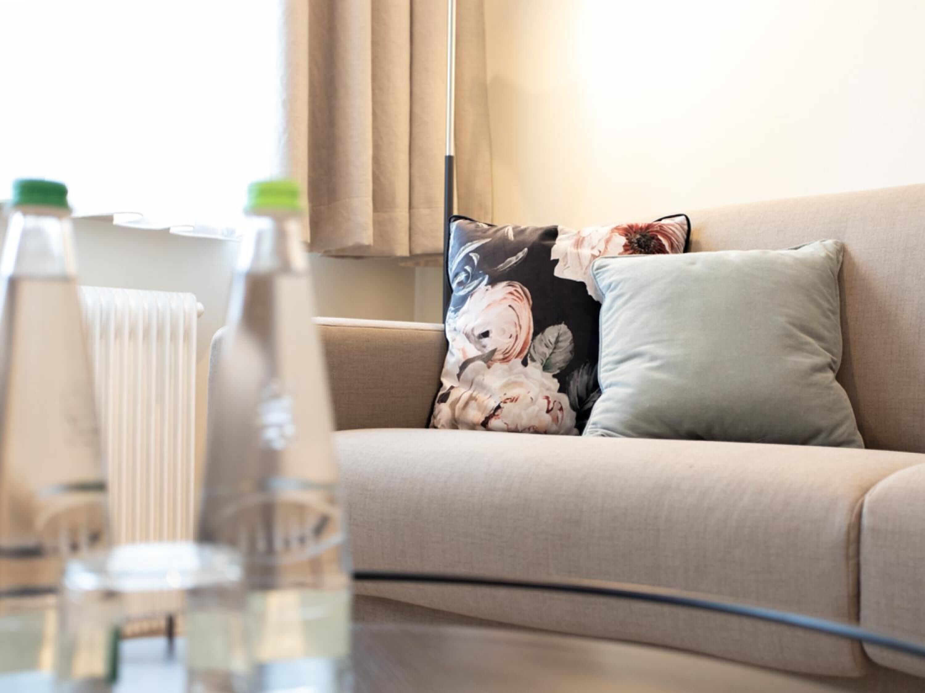 En stue med en beige sofa, blomstermønstrede puder og et sofabord med flasker.