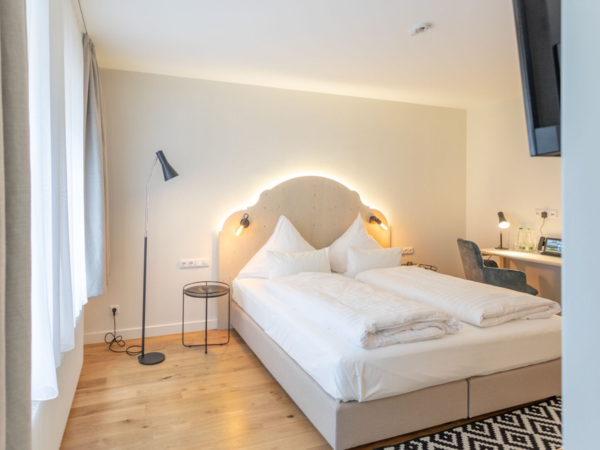 A modern hotel room with a large bed, bedside lamps, a desk with a chair, and a view of the city.