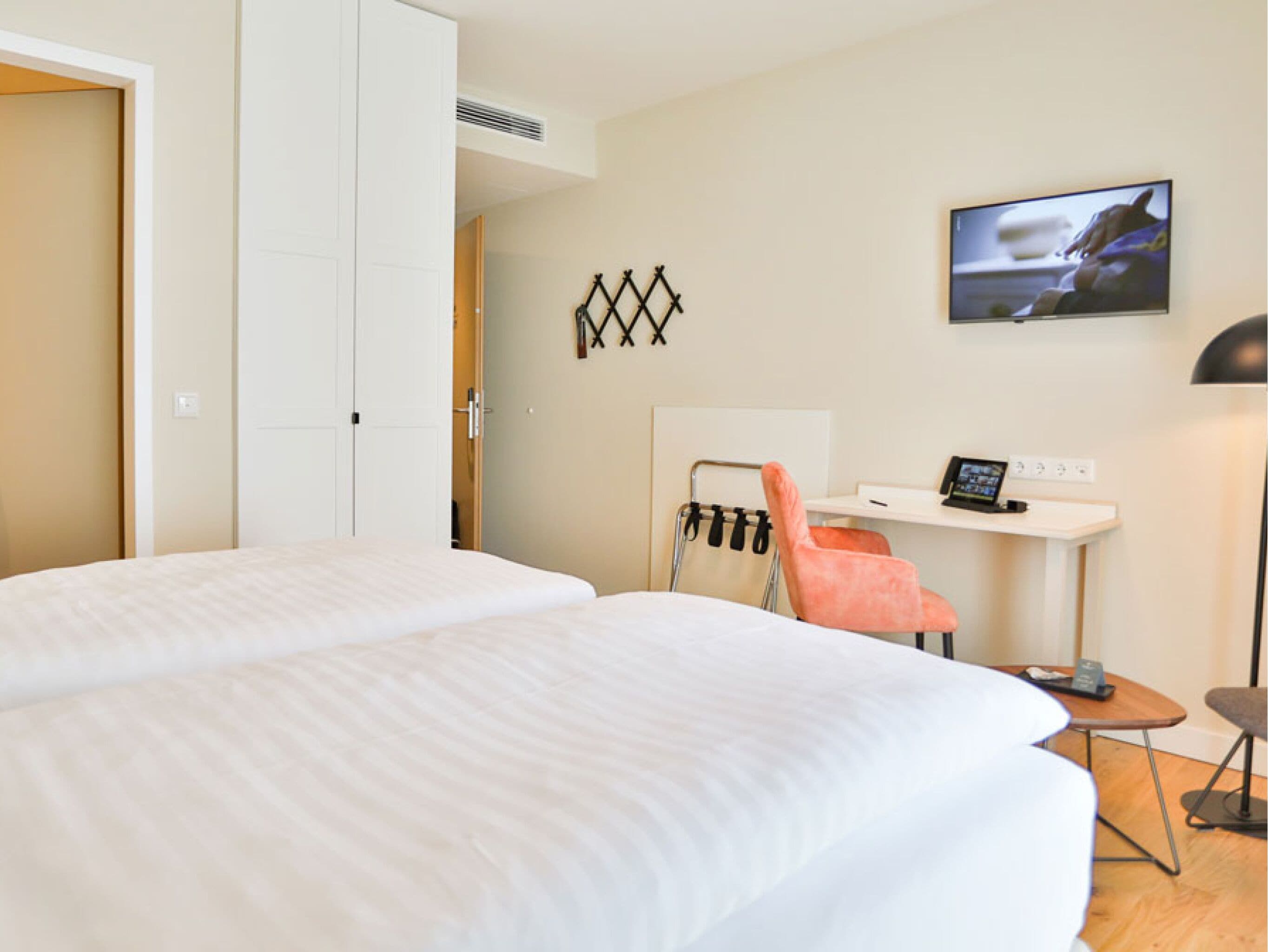 A hotel room with two beds, a desk with a computer, a chair, and a TV on the wall.