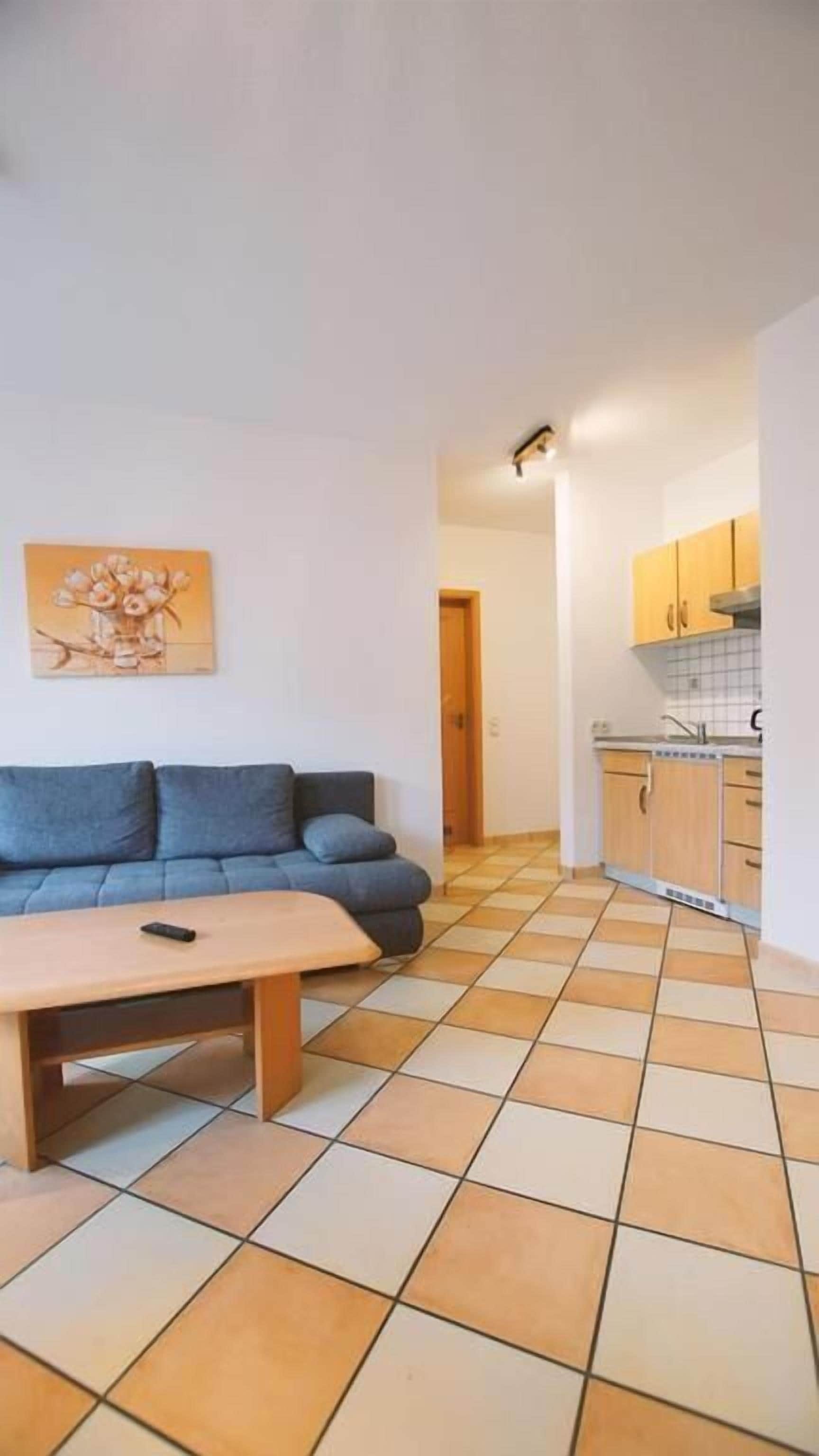 Una sala de estar con un sofá y una mesa de centro, una cocina con armarios y un lavamanos, y un piso con baldosas.