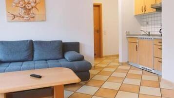 A living room with a sofa and a coffee table, a kitchen with cabinets and a sink, and a tiled floor.